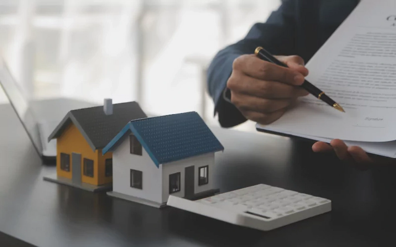 Real estate contract signing with house models, calculator, and property purchase documents on a desk.